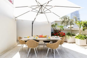 An outdoor dining area with a white umbrella, wicker chairs, and a table set with plates, bowls, and fruits.