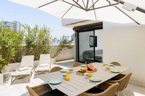A rooftop terrace with a dining table set for four, surrounded by wicker chairs, a white umbrella, and a city view. - Hippie Chic at Hovevei with MAMAD by HolyGuest (Tel Aviv)