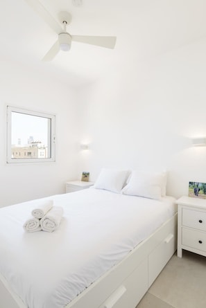A white hotel room with a bed, a nightstand, a bedside table, a ceiling fan, and a window with a city view.