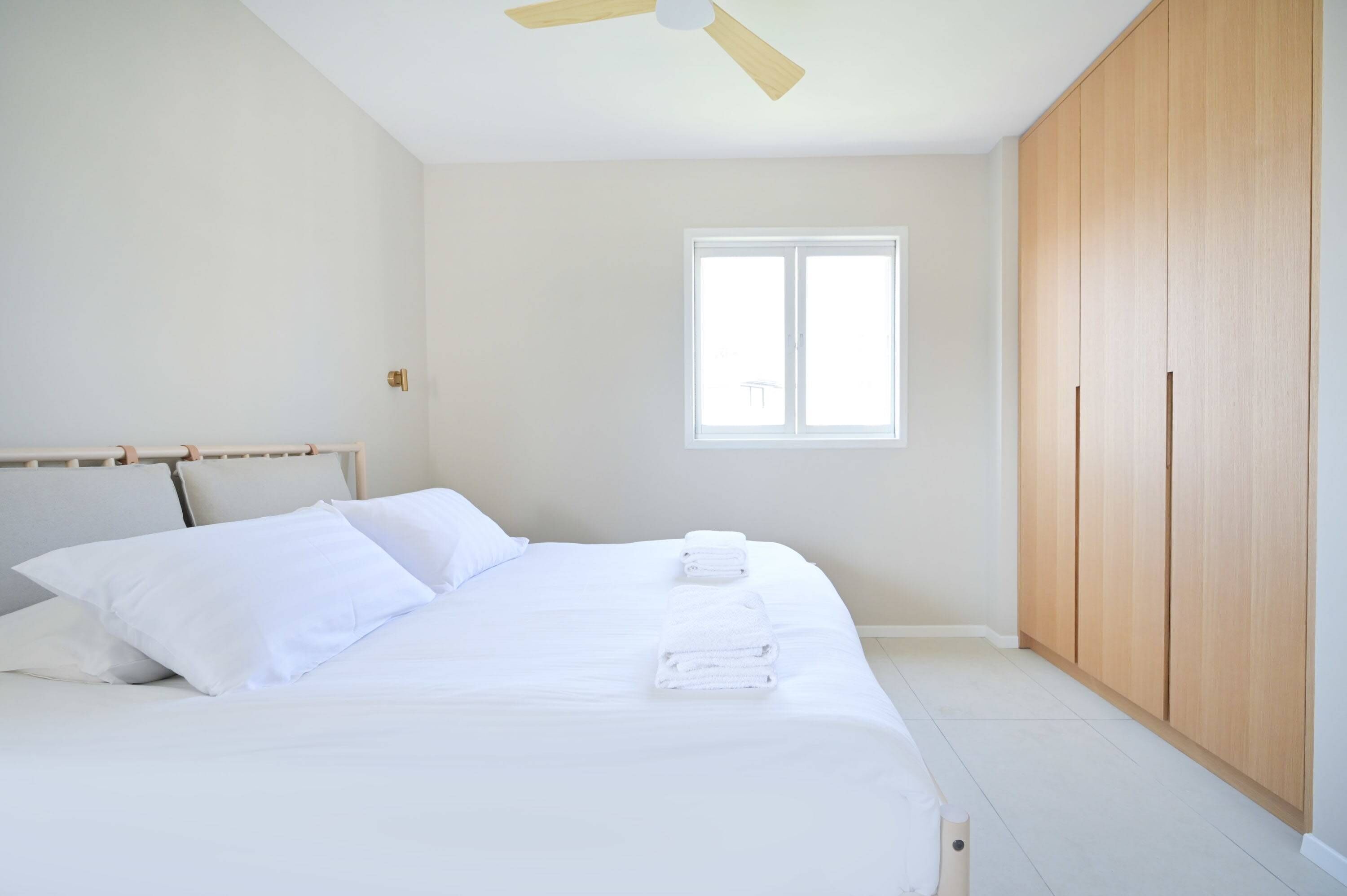 A bedroom with a bed, white linens, a window, a ceiling fan, and wooden wardrobe.