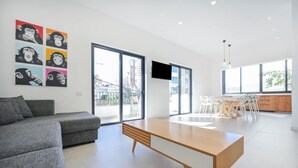 Ein modernes Wohnzimmer mit einer grauen Couch, einem Holztisch und einem Essbereich mit weißen Stühlen. Große Fenster sorgen für natürliches Licht und bieten einen Blick auf die Stadt.