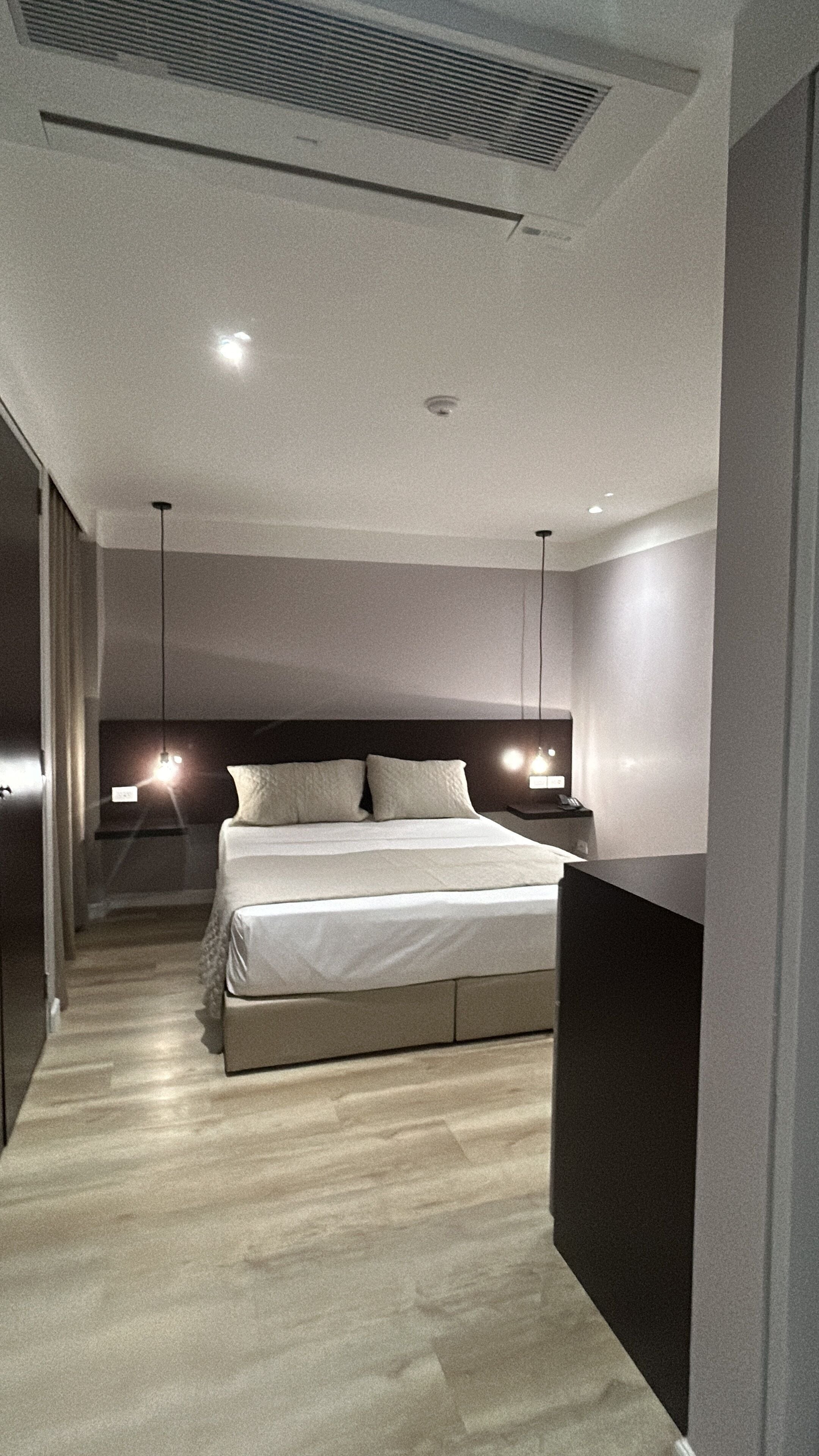 A modern hotel room with a large bed, bedside tables, a ceiling vent, and a hallway leading to another room.