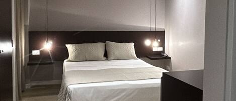 A modern hotel room with a large bed, bedside tables, a ceiling vent, and a hallway leading to another room.