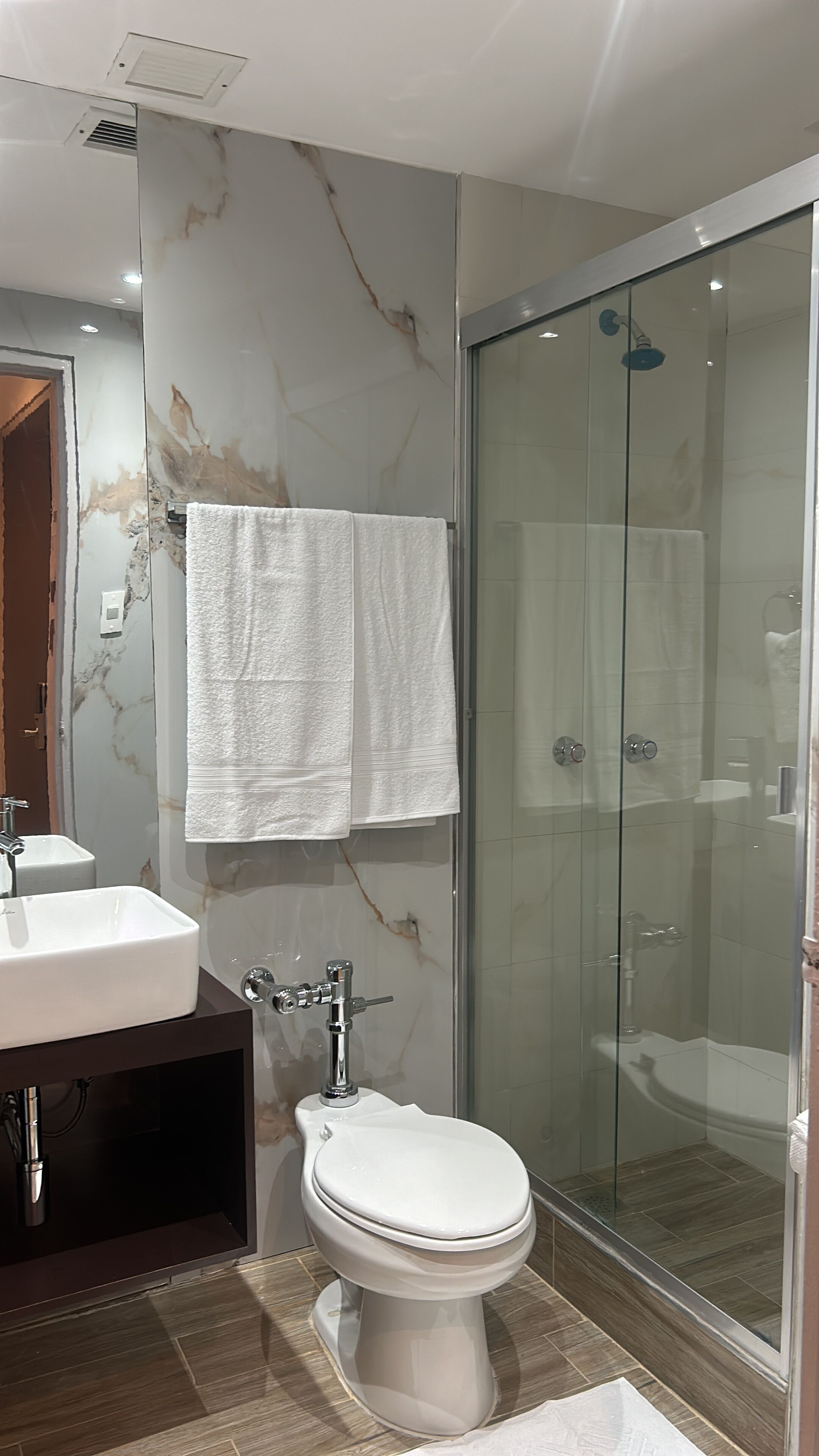 Un baño con pared de mármol, cabina de ducha de vidrio, inodoro blanco y lavamanos con un mueble oscuro.