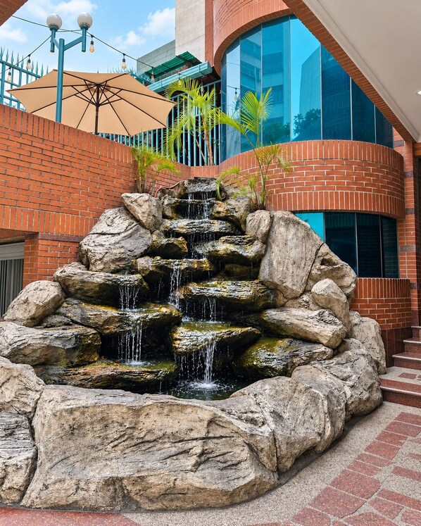 Une cascade artificielle en roche, aménagée dans une cour extérieure, avec un bâtiment moderne en arrière-plan.