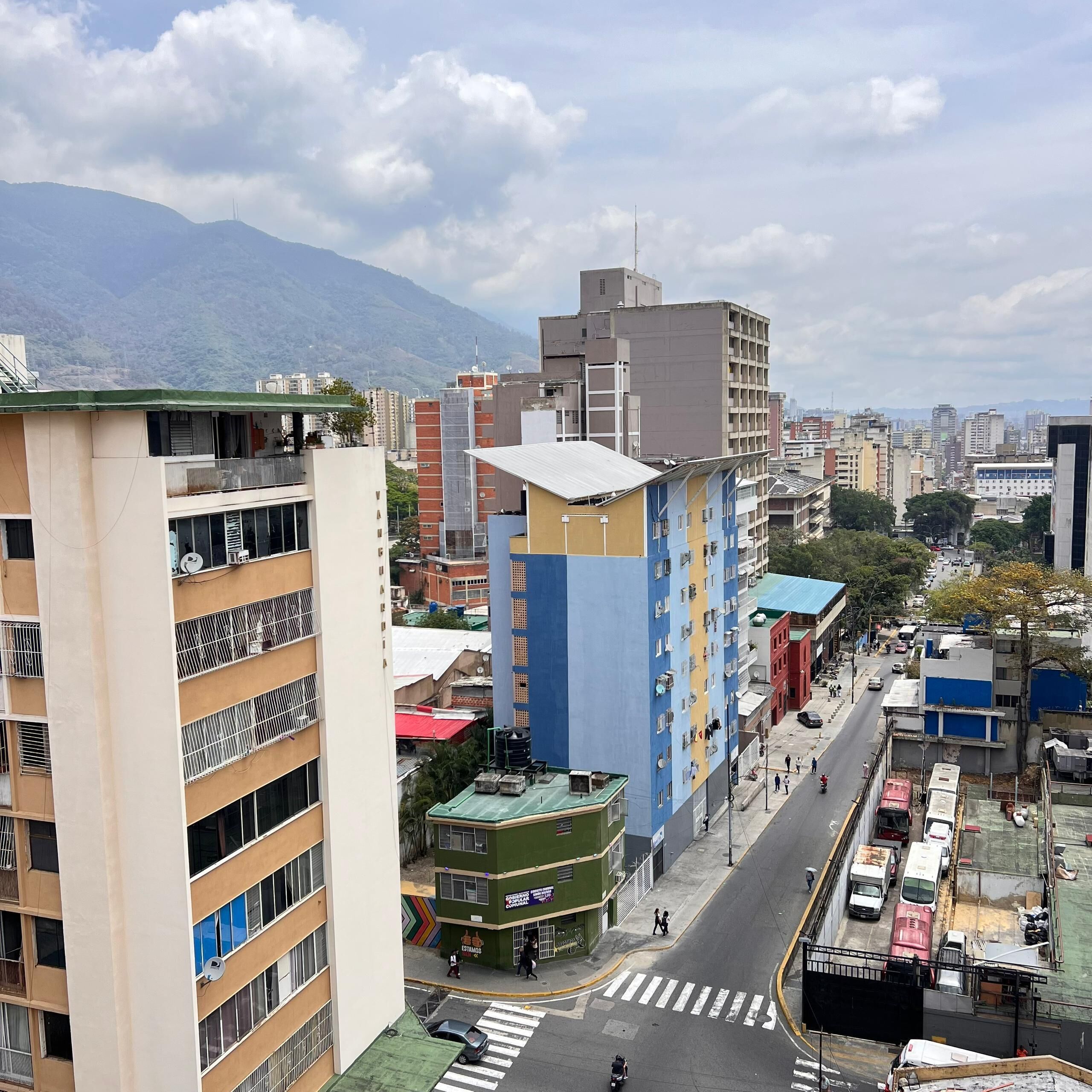 Un edificio de varios pisos con balcones, una calle con vehículos y una montaña al fondo.