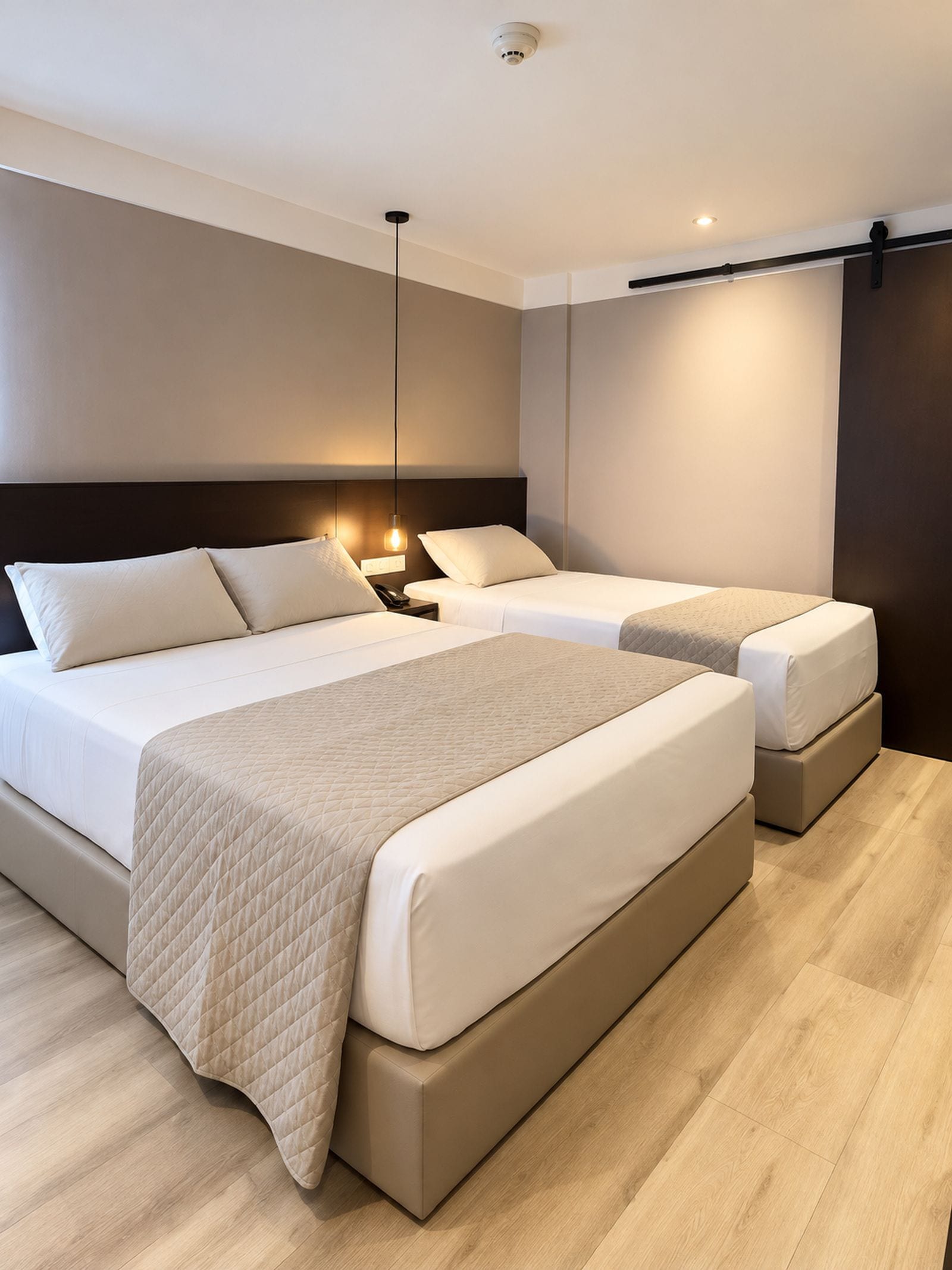 A modern hotel room with a large bed, two smaller beds, a bedside table with a lamp, and a dark-colored headboard.