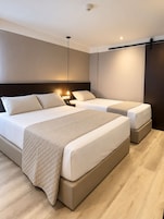 Una habitación de hotel moderna con una cama grande, dos camas más pequeñas, una mesita de noche con lámpara y un cabecero oscuro.