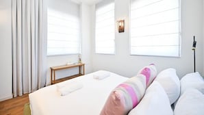 Habitación de hotel con una cama grande, cortinas blancas, una mesita de noche con lámpara y una ventana con persianas.