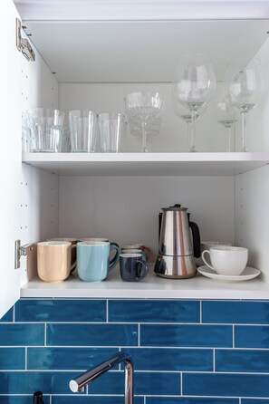 A kitchen with a white cabinet shelf holding clear wine glasses and a stainless steel coffee maker. Below, there are various ceramic mugs and cups. - Red Brick Apartament w Rynku ProperUNIT (Wroclaw)