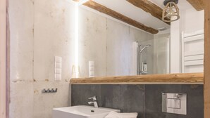 A bathroom with a white sink, a wooden stool with towels, a mirror, and a shower. - Red Brick Apartament w Rynku ProperUNIT (Wroclaw)