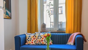 A blue sofa with patterned cushions, a small round wooden table with a vase of flowers, and a view of buildings through a window with yellow curtains. - Red Brick Apartament w Rynku ProperUNIT (Wroclaw)