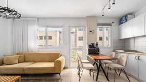 Una sala moderna con sofá, mesa de comedor y sillas, una cocina con armarios y electrodomésticos, y ventanas amplias con vistas a edificios.