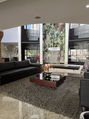 Um lobby moderno com um sofá preto, uma mesa de centro de vidro com um vaso, e uma grande janela com vista para a rua.