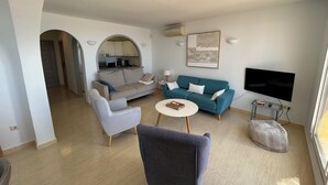 Een moderne woonkamer met een grijze bank, een blauwgroene bank, een grijze fauteuil en een witte salontafel. Er is een flatscreen televisie aan de muur bevestigd, een klein bijzettafeltje en een kruikzak stoel.