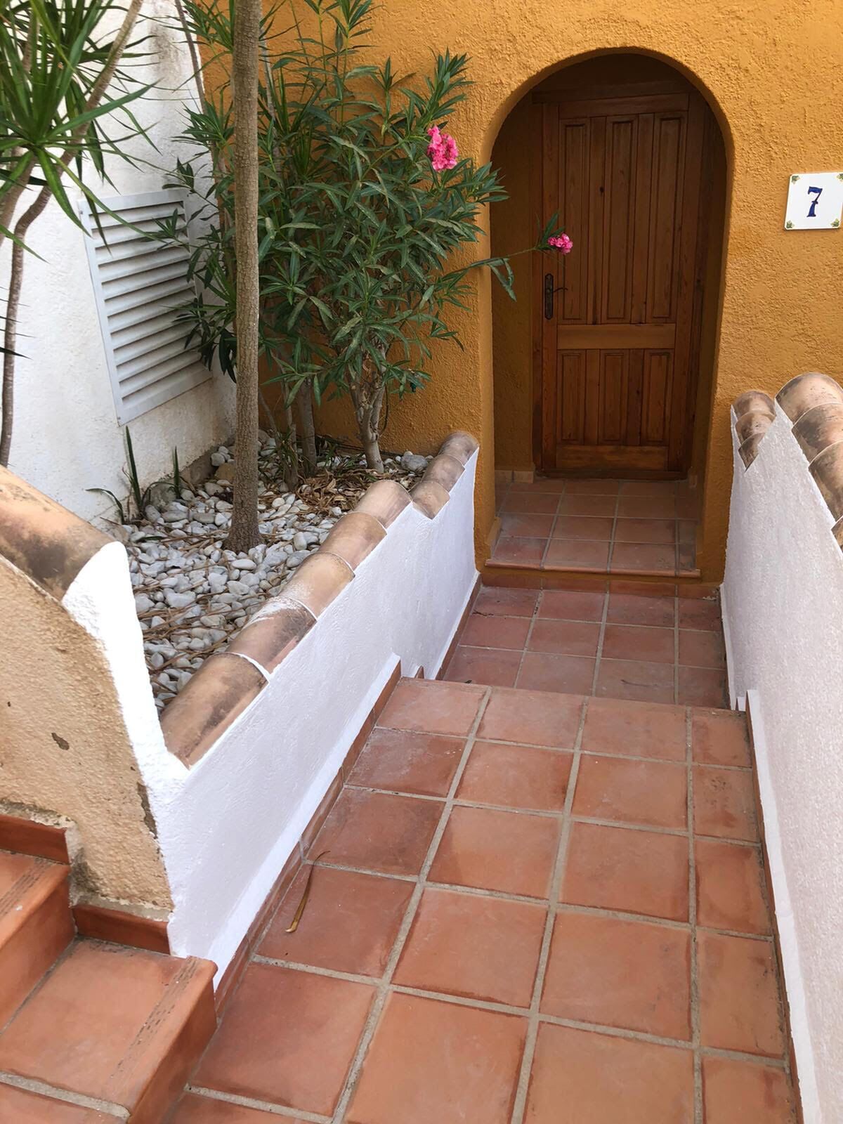 A narrow, tiled corridor with a wooden door and a white number '7' sign, leading to a building with a yellow wall and a small tree with pink flowers.