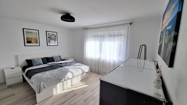 Une chambre moderne avec un lit, des tables de chevet, un bureau et une grande fenêtre ornée de rideaux légers.
