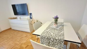 Une petite pièce propre, avec un plancher en bois, un bureau blanc recouvert d’une nappe à motifs floraux, une plante en pot et un téléviseur à écran plat posé sur une commode blanche.