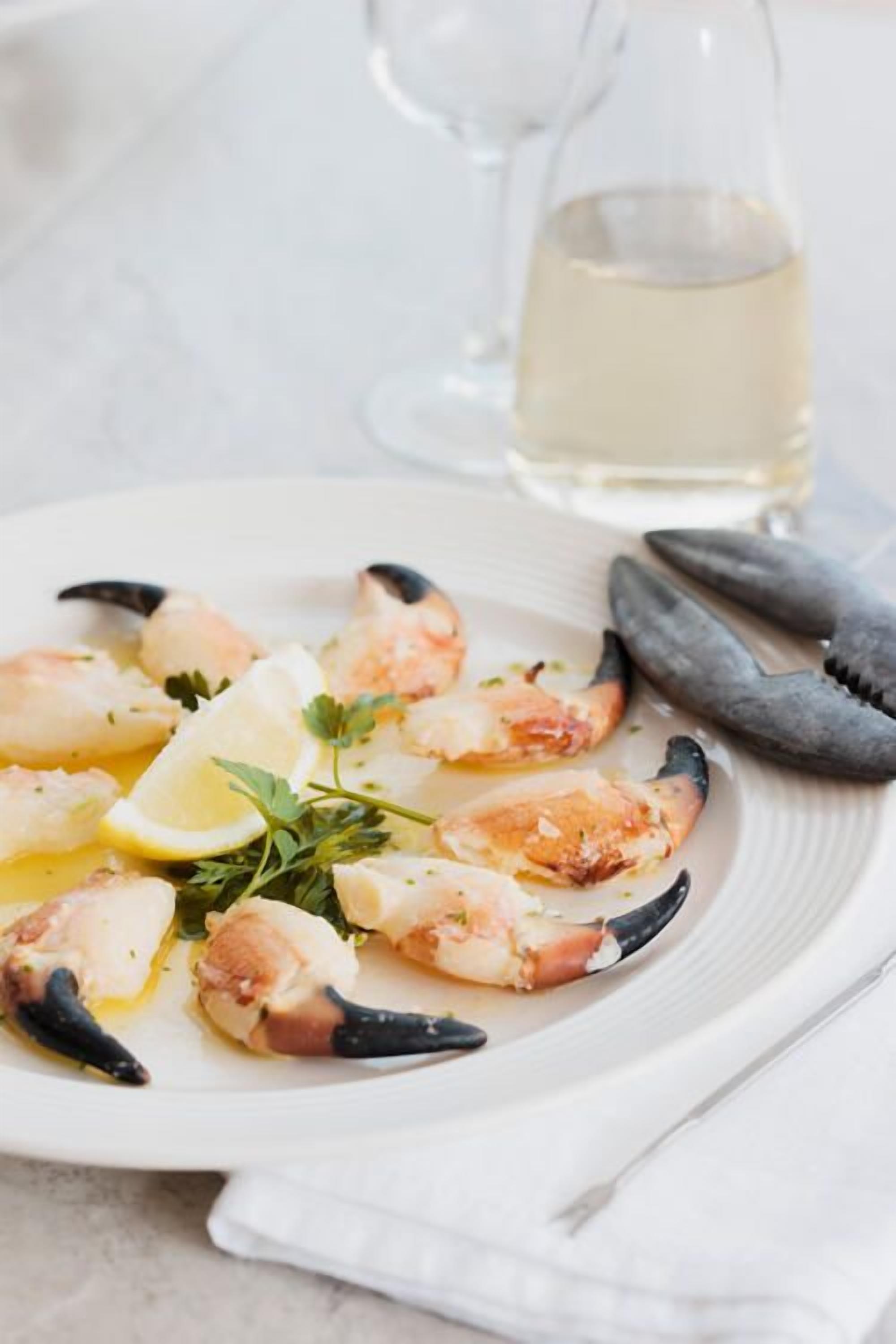 A plate of cooked seafood, likely shrimp, garnished with lemon and herbs, accompanied by a glass of white wine and a pair of tongs.