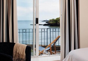A room with a large window overlooking a coastal view, a bed, a chair, and a small table with a bottle.