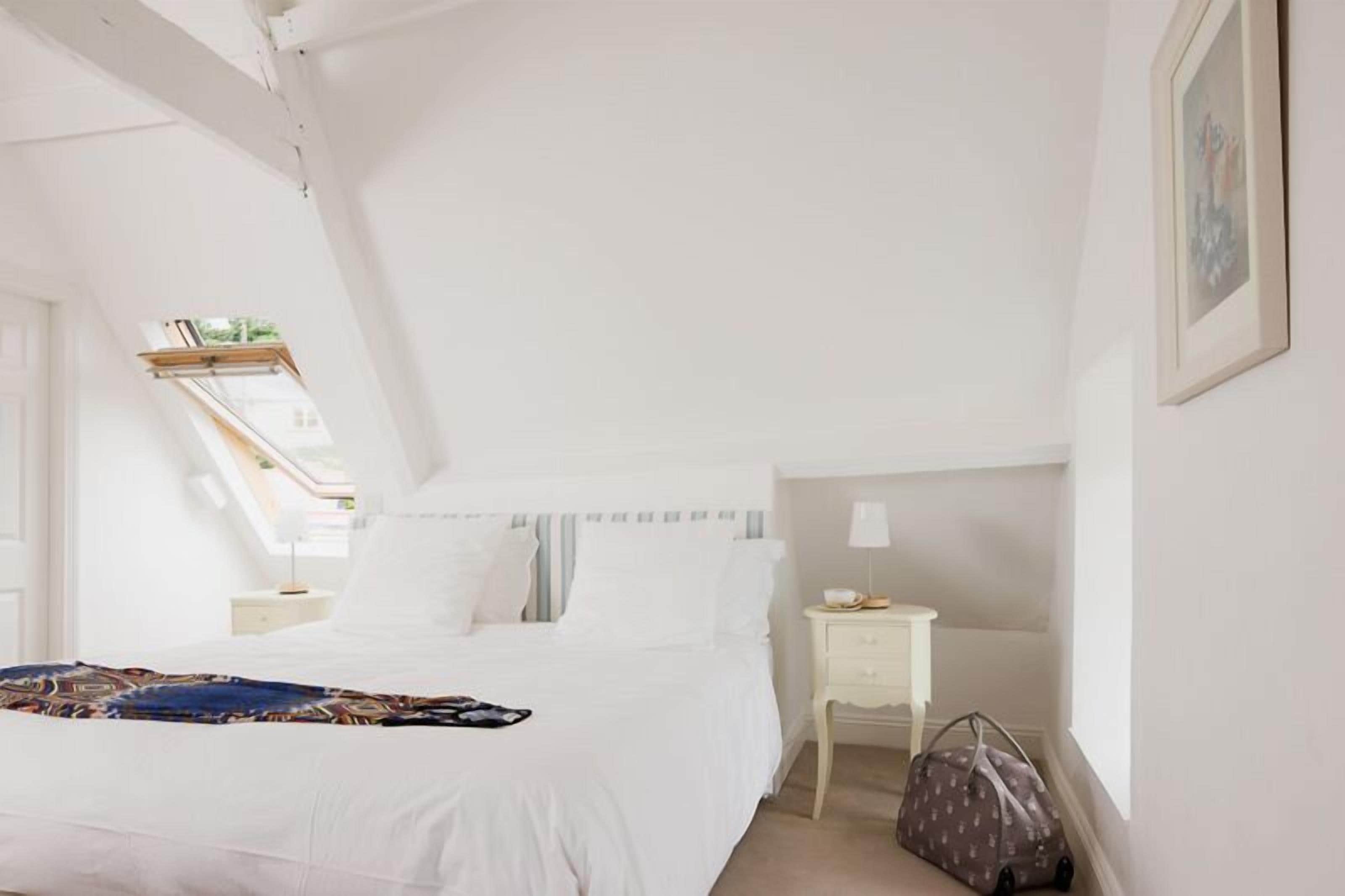 A bedroom with a bed, a bedside table, a lamp, a bag, and a window with a view of greenery.