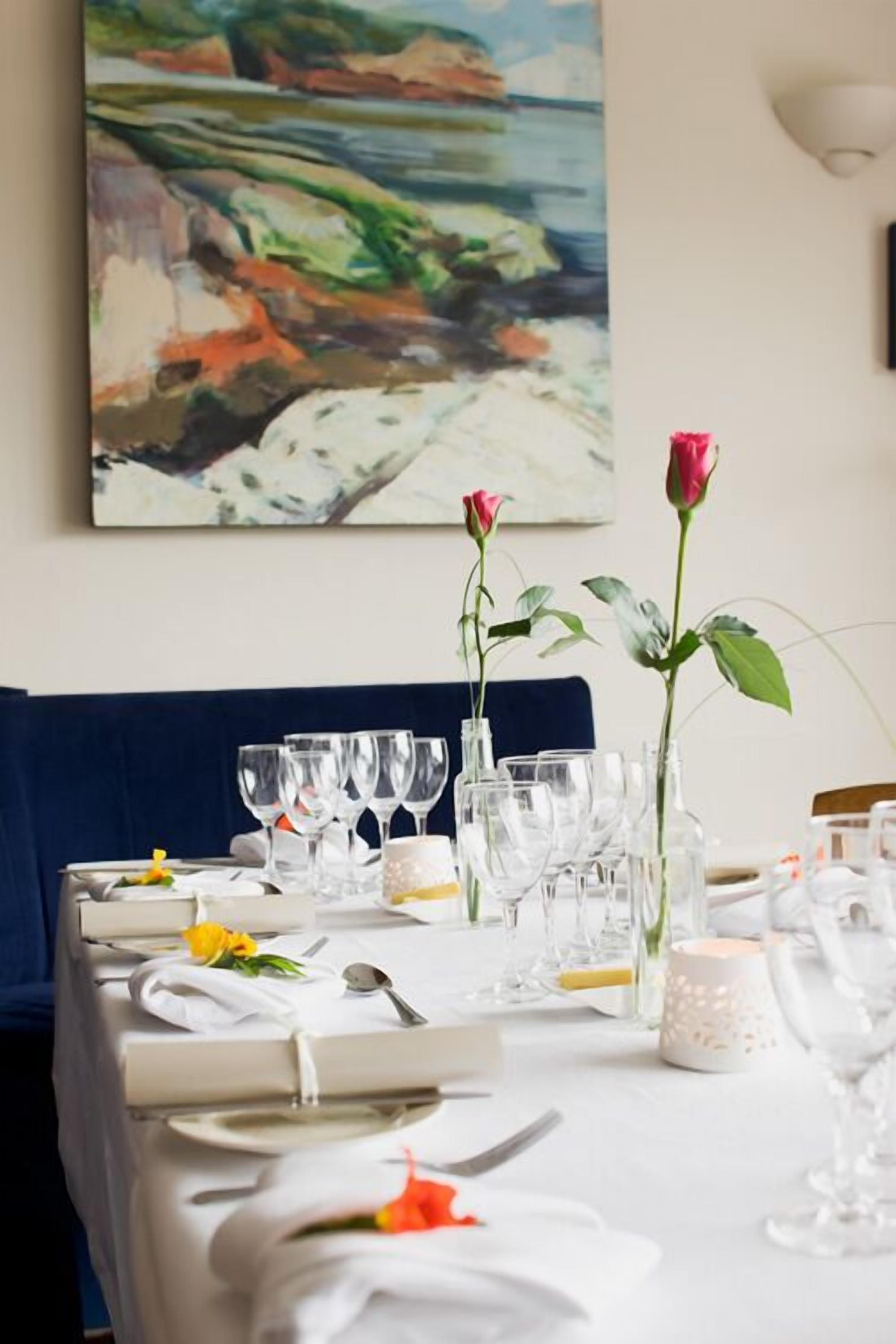 Une table à manger dressée avec une nappe blanche, de la vaisselle transparente et un tableau représentant un paysage accroché au mur.