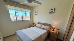 Un dormitorio con cama, mesitas de noche, ventilador de techo, ventana con vistas y una obra de arte en la pared.
