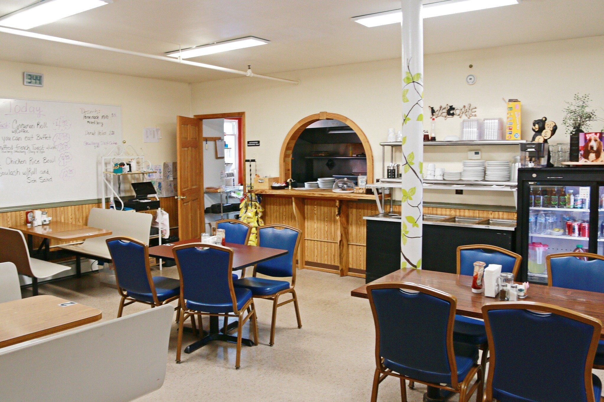 A dining area with tables and chairs, a whiteboard with notes, a kitchen area with a refrigerator, and a shelf with various items.