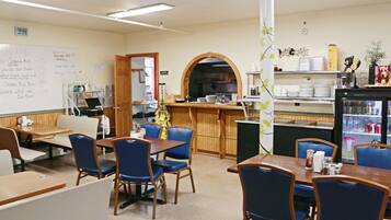 A dining area with tables and chairs, a whiteboard with notes, a kitchen area with a refrigerator, and a shelf with various items.