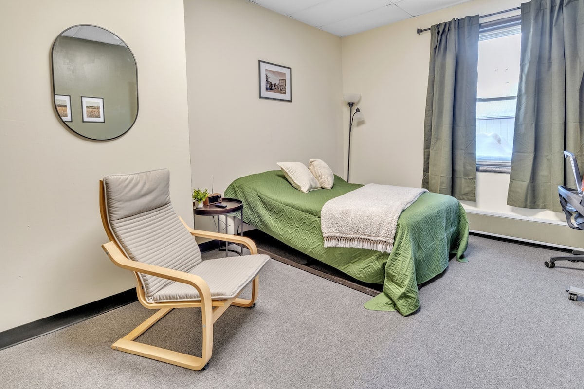 Un dormitorio con cama, una silla, una mesita de noche, una lámpara y una ventana con cortinas.