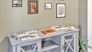 A room with a white cabinet displaying magazines and a notebook, a potted plant, and framed pictures on the wall. - Foothills Lodge & Diner (Malone)