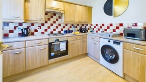 A compact kitchen with wooden cabinets, a built-in oven, a washing machine, and a microwave. - Spacious House | Parking | Business & Leisure (Gloucestershire)