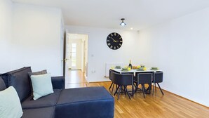 A modern living room with a dark blue sofa, a dining table with chairs, and a wall clock.