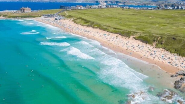 Una playa costera con aguas turquesas cristalinas, una orilla arenosa y un área con césped con edificios al fondo.