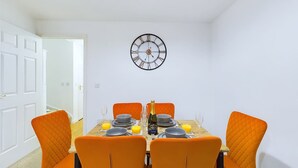 A dining room with a table set for four, featuring orange chairs and a wall clock.