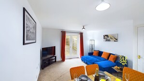 A living room with a blue sofa, orange chairs, a dining table set, a TV, and a door leading outside.