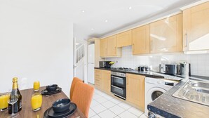 A modern kitchen with wooden cabinets, a built-in oven, a washing machine, and a dining area with a table set for two. - *NEW* Prestbury Place | Racecourse | Free Parking (Prestbury)