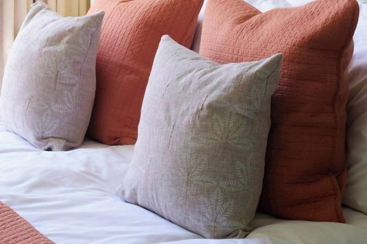 Una cama con almohadas blancas y color melocotón, una lámpara color beige y un papel tapiz con estampado de palmeras.