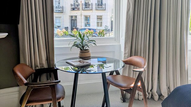 Ett modernt hotellrum med ett glasbord, två läderfåtöljer, en krukväxt och utsikt över en vit byggnad med balkonger.