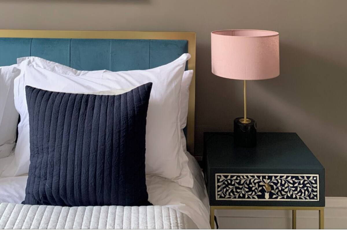 Una cama bien tendida con un cabecero azul, una lámpara rosa en una mesita de noche y una obra de arte colorida en la pared.
