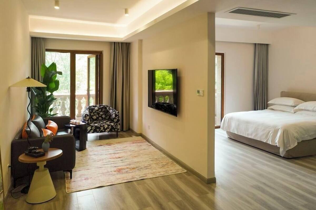 Una habitación de hotel moderna con una cama, un televisor montado en la pared, una zona de estar con una mesa y sillas, y un ventanal con cortinas.