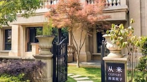 Un edificio de dos pisos con estilo arquitectónico clásico, que presenta una entrada central con una reja negra, un pequeño jardín con losetas y un letrero con texto en un idioma extranjero.