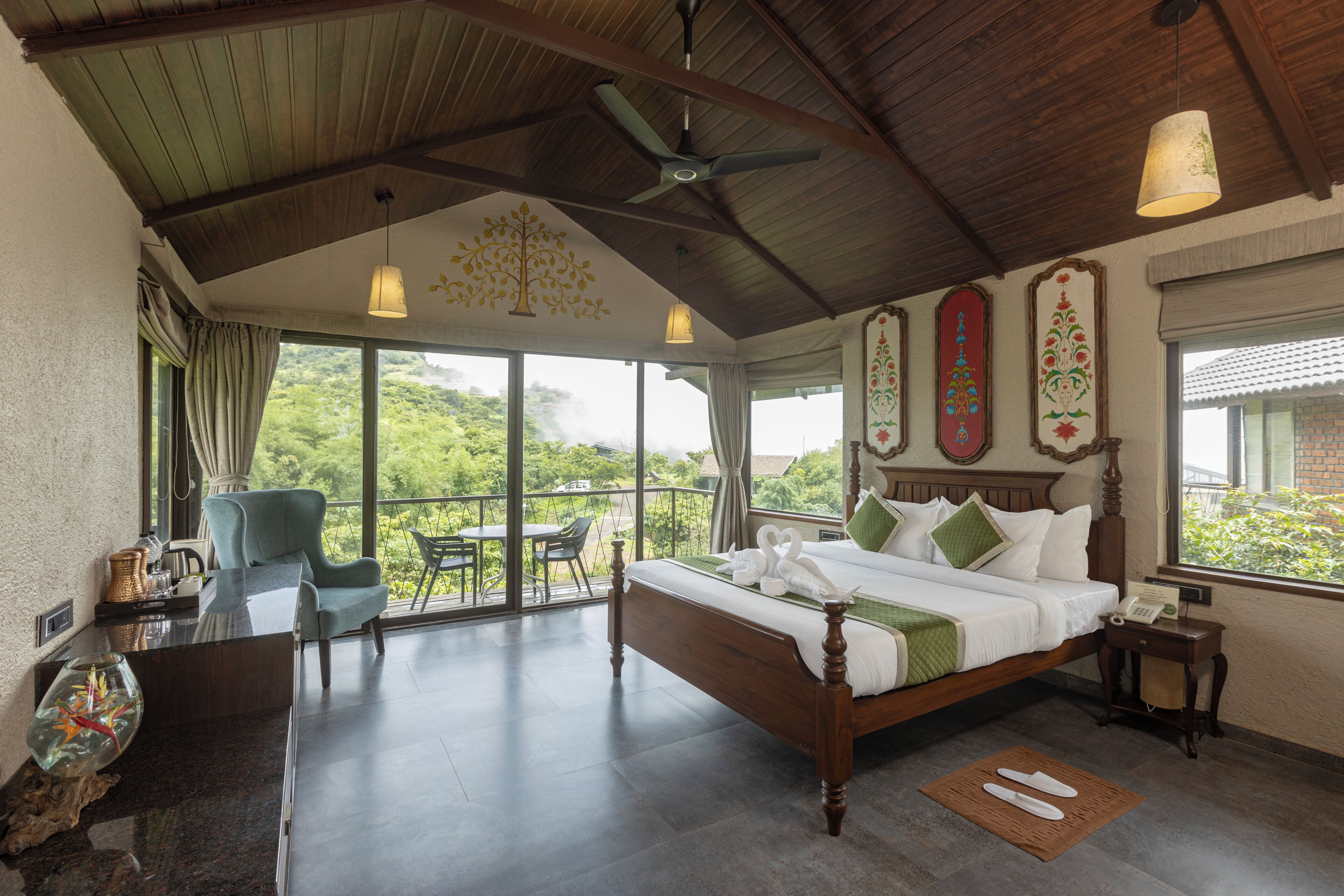 A spacious bedroom with a large bed, a sitting area with a chair and table, and a balcony with a view of greenery.