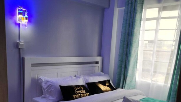 Un lit bien fait, avec une literie blanche et un plaid turquoise, une table de chevet avec une lampe, et une fenêtre avec des rideaux.