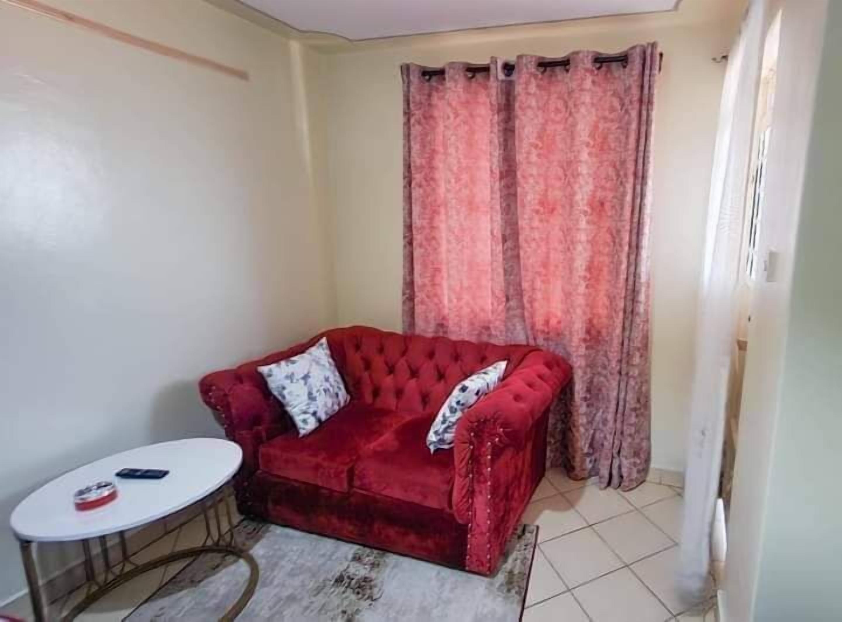 Un sofá de terciopelo rojo con un cojín estampado, una mesita redonda con un control remoto, y una ventana con cortinas rosadas.