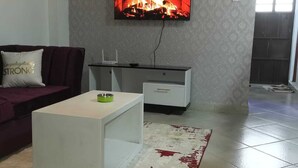 Un salon avec un téléviseur fixé au mur qui affiche une image de cheminée, un canapé avec un coussin, une table basse et un tapis à motifs.