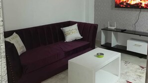 Un salon avec un canapé violet, une table basse blanche, un téléviseur à écran plat fixé au mur et un tableau au-dessus du canapé.
