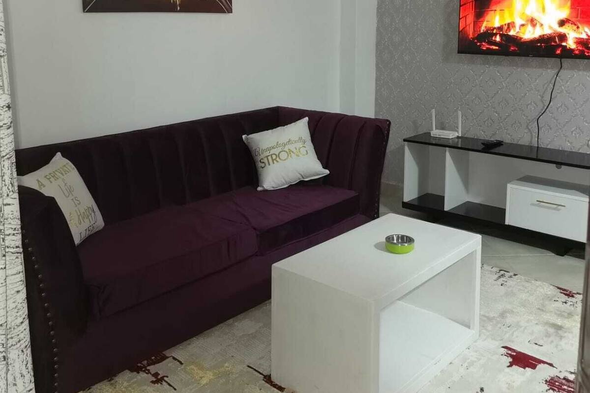 A living room with a purple sofa, a white coffee table, a flat-screen TV mounted on the wall, and a painting above the sofa.
