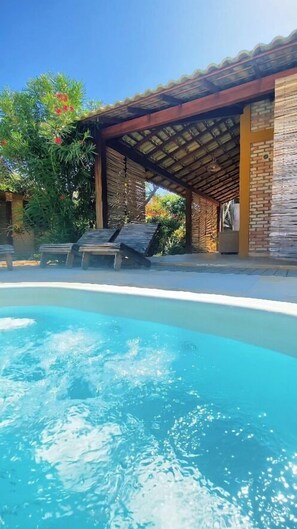 A pool area with a clear blue pool, wooden lounge chairs, and a covered patio with a tiled roof. - Vila Flamboyant (Cajueiro da Praia)