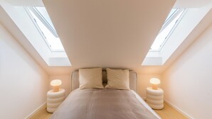 Un dormitorio con tragaluces, una cama con colcha color beige, dos lámparas de noche y suelo de madera.